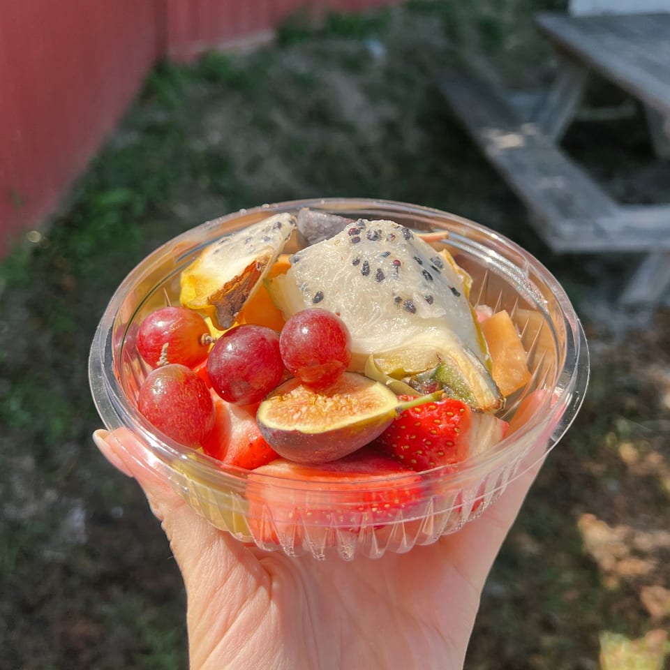A hand holds a clear plastic container filled with fresh fruit, including grapes, figs, strawberries, and dragon fruit.