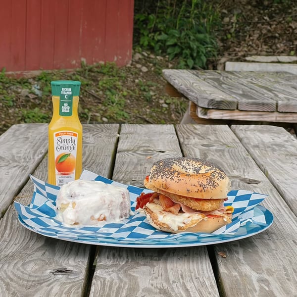 A Country Corners Deli bagel sandwich, cinnamon roll, and orange juice on a wooden picnic table.