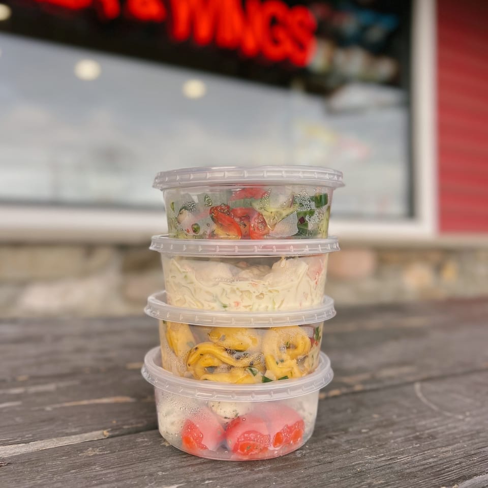 Stack of four clear plastic containers filled with various deli salads on a wooden surface.