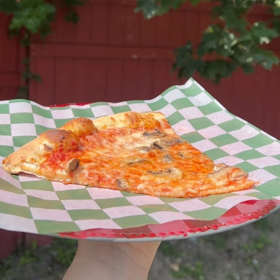 A slice of mushroom pizza on green and white checkered paper, held against a red wall.