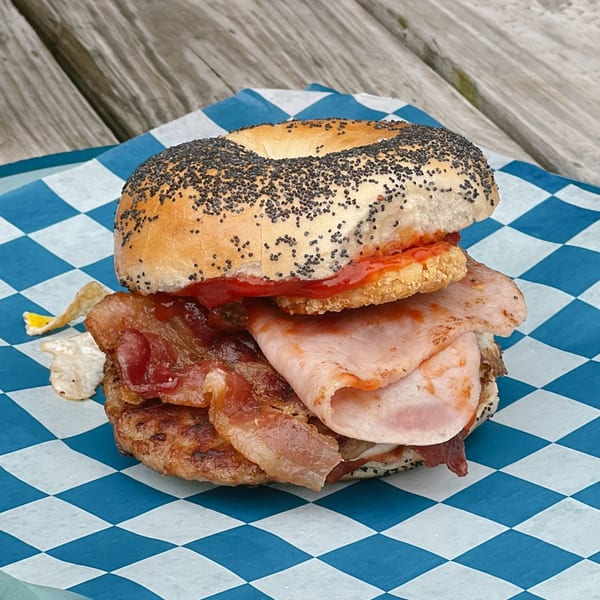 Poppy seed bagel with bacon, ham, and sauce on blue and white checkered paper.