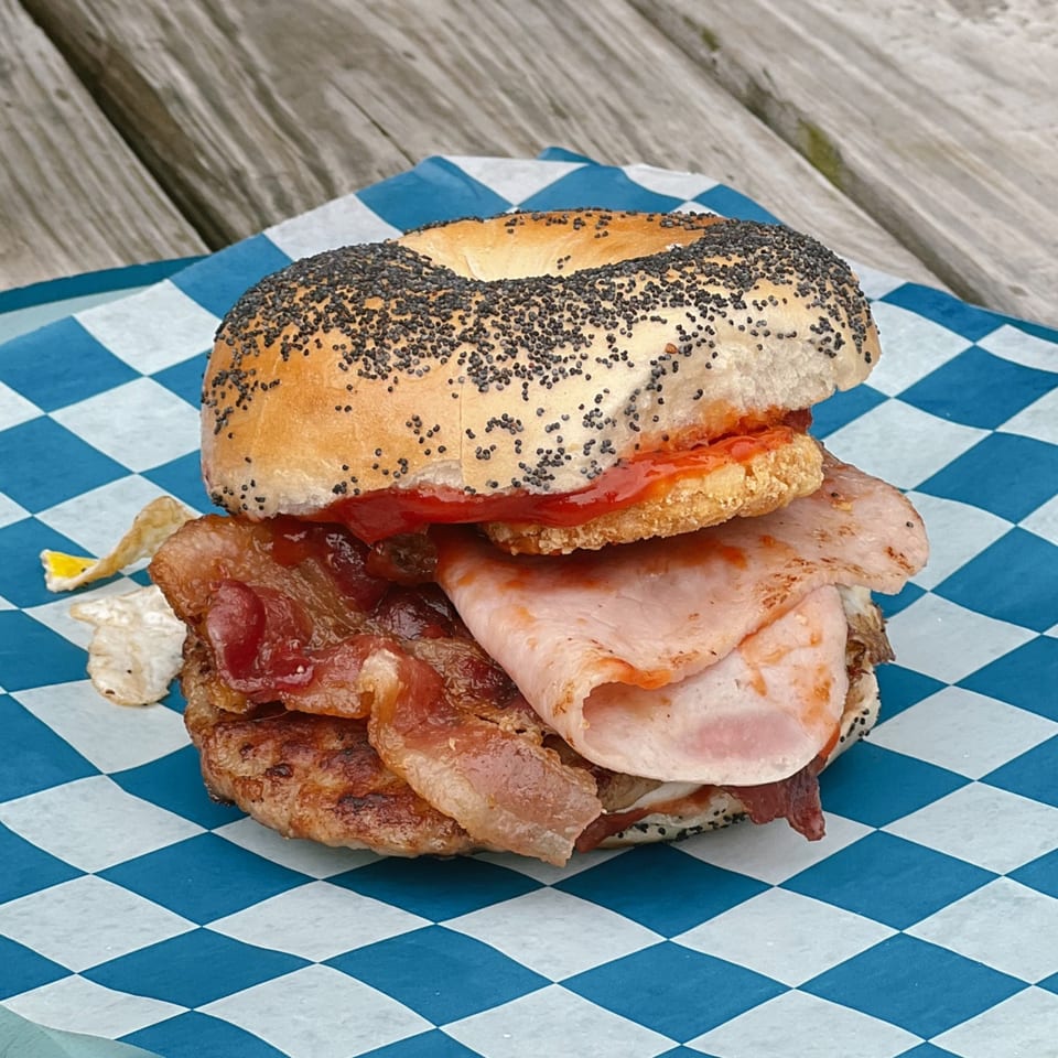 Poppy seed bagel with bacon, ham, and sauce on blue and white checkered paper.