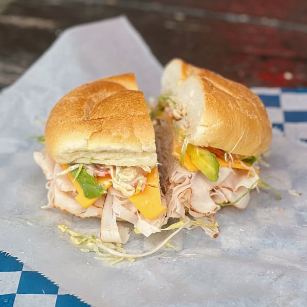 A Country Corners Deli sandwich, cut in half, with turkey, cheese, avocado, and lettuce on a roll.